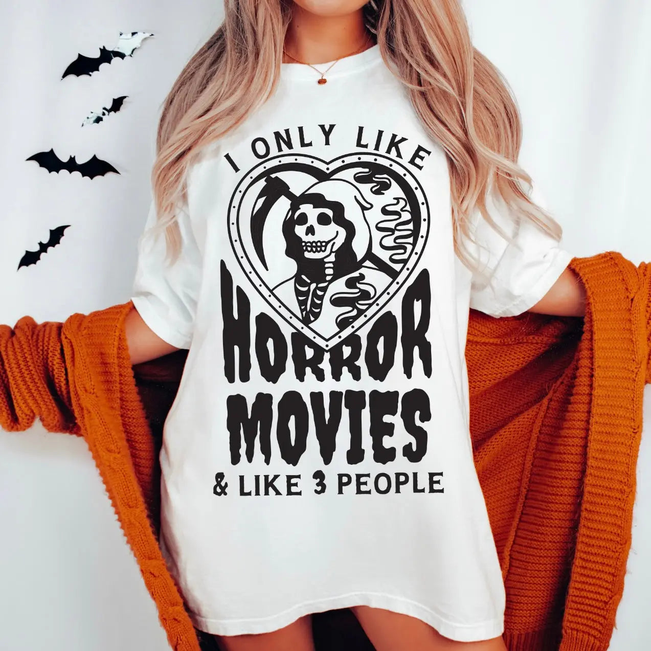 Woman wearing the Horror Movies • Tee, a white unisex airlume combed cotton t-shirt with a skeleton and heart graphic, reading I only like horror movies & like 3 people, holding an orange cardigan with black bat wall decor behind her.