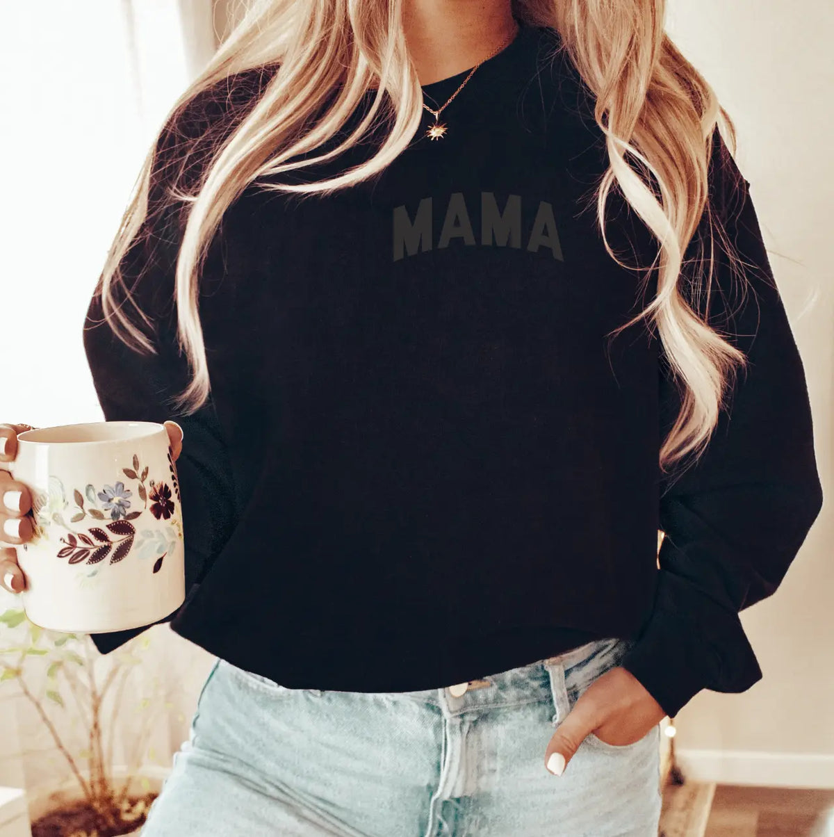 A woman with long, wavy blonde hair wears the MAMA • Embossed Black Pullover and holds a floral mug by a window. Her face isn’t visible. The unisex sweatshirt offers cozy comfort and is paired here with casual blue jeans.