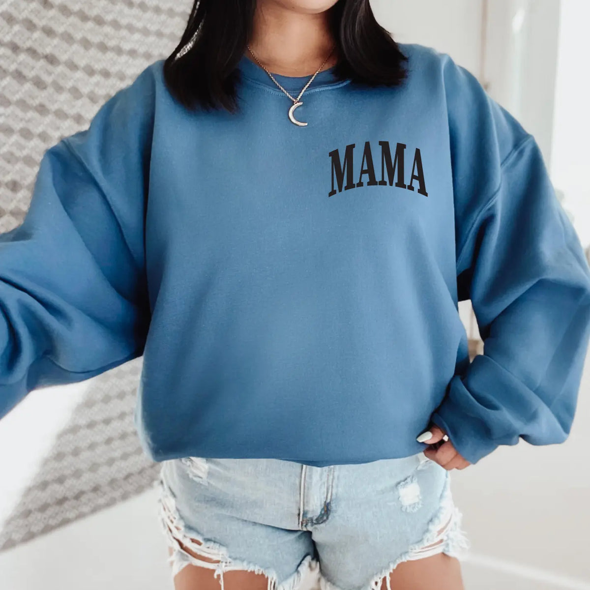 A woman stands indoors wearing the MAMA Arch Chest • Indigo Pullover, paired with light blue ripped denim shorts and a crescent moon necklace. Her face is not visible.