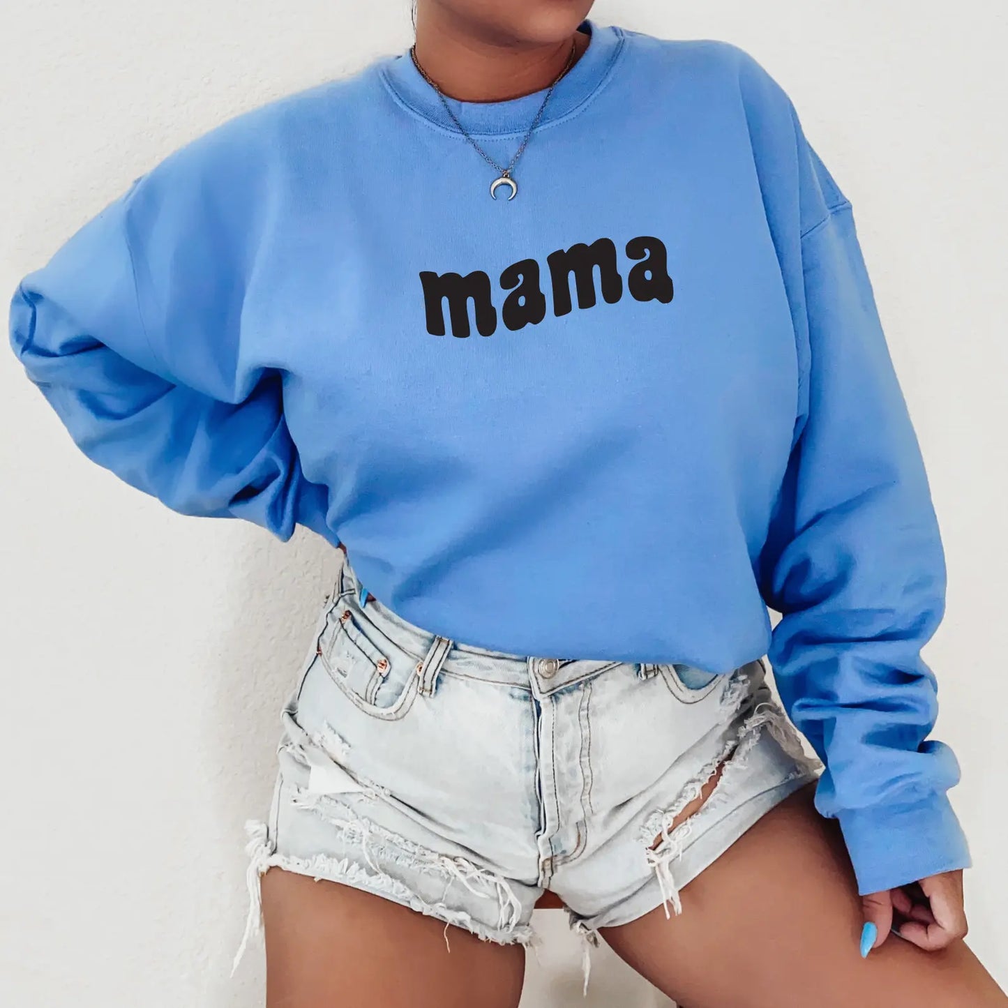 Wearing the Mama Wave • Carolina Pullover, a unisex blue sweatshirt with mama in black letters, and light denim ripped shorts, this look is perfect for cooler weather against a plain light background.