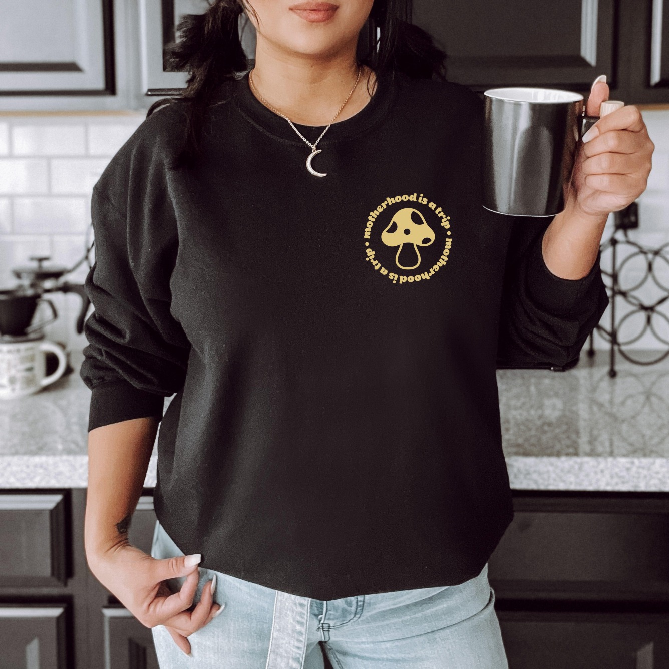 In a kitchen, a woman in pigtails wears the “Motherhood is a Trip” Black Pullover with a yellow mushroom graphic, paired with a crescent moon necklace and holding a black mug.