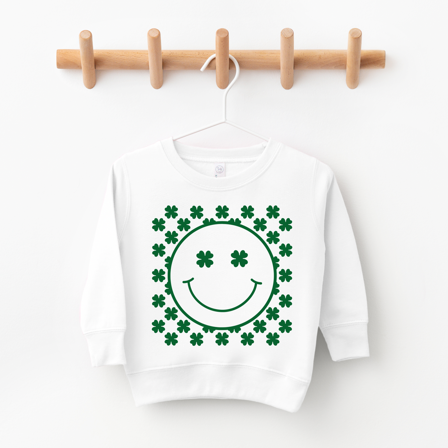 The Happy Shamrock white kids sweatshirt features a green smiley face with clover eyes, surrounded by clovers. Unisex fit—great for sizing up as your toddler grows. Displayed on a hanger and wooden wall hook.
