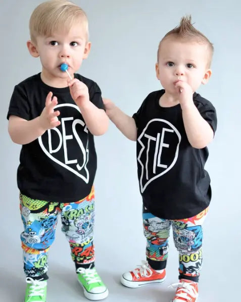 Two toddlers stand side by side, each holding a blue lollipop. They wear matching Bestie • Black Tees with “BEST” and “TIE” heart designs, comic book-print pants, and green and red sneakers—playful unisex comfort.