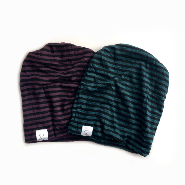 Two Striped Beanies, one in dark purple/black and the other in dark green/black, feature horizontal stripes and a small white logo tag at the edge. Made of comfortable interlock knit for a stylish look, shown on a white background.