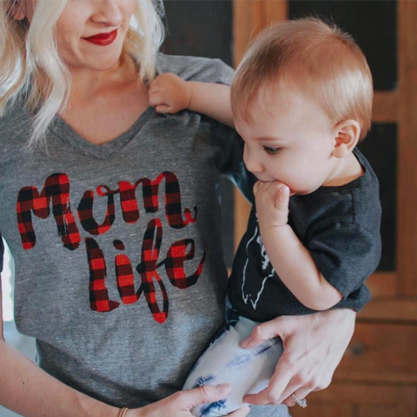 A woman wearing a gray Mom Life Plaid • Tee holds a baby dressed in a dark shirt and buffalo plaid pants. The baby, with light hair and a hand in their mouth, is partially visible next to her indoors.