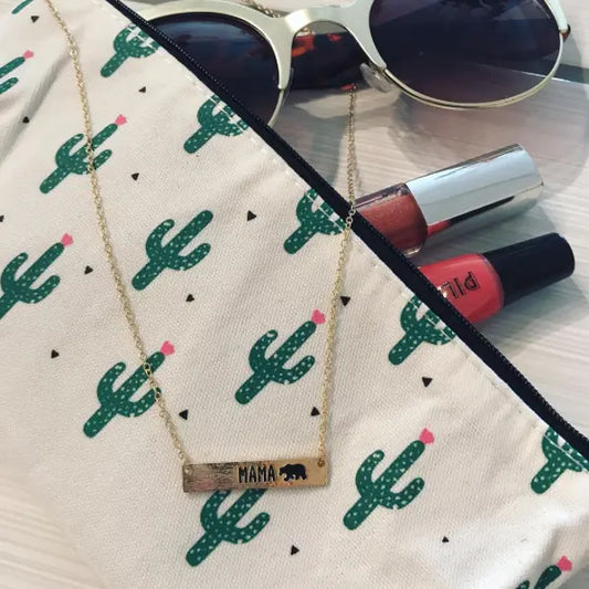The Mama Bear • Bar Necklace, available in gold or silver with an adjustable clasp and bear charm, rests on a cactus-patterned pouch beside sunglasses and two lip gloss tubes.