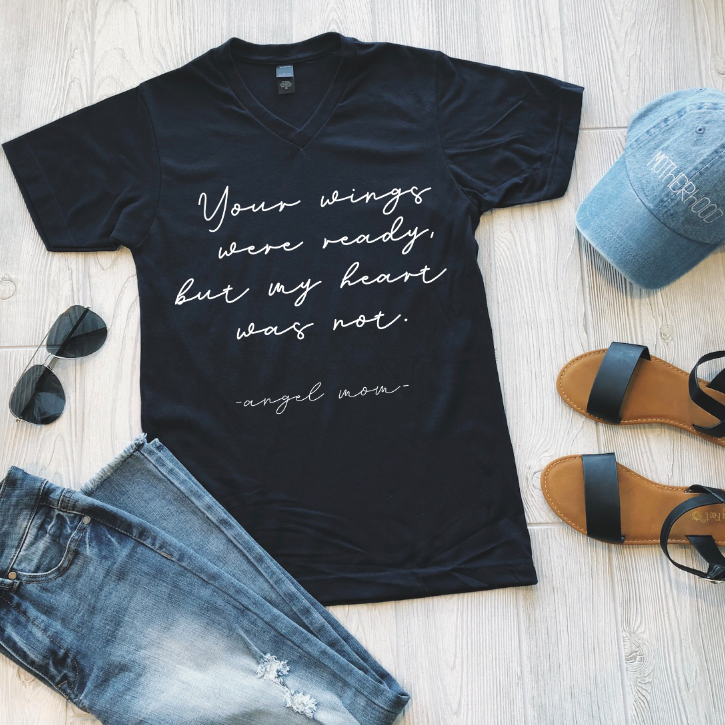 A flat lay of the Angel Mom • Navy Tee, featuring white script “Your wings were ready, but my heart was not.” Crafted from soft ring-spun cotton, its styled with jeans, sandals, a blue hat, and black sunglasses on a wooden floor.