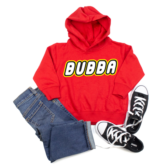 The Bubba • Lego Hoodie, a true-to-size red hoodie with BUBBA in bold yellow and white lettering, is styled with blue jeans and black lace-up sneakers with white soles—displayed flat on a white background.