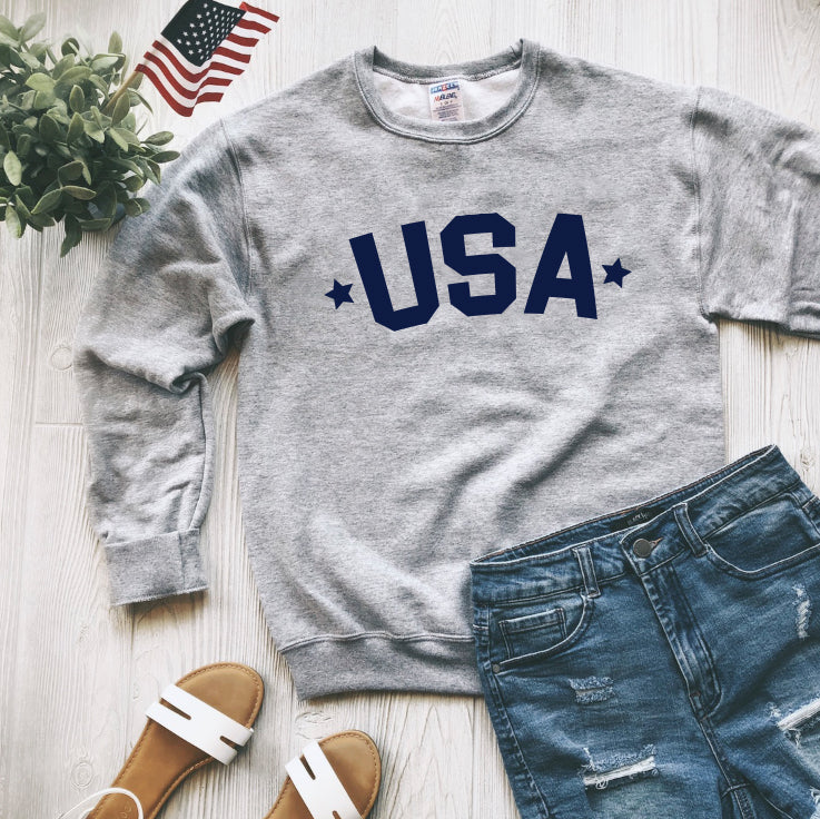The USA • Adult Pullover, a gray unisex sweatshirt with navy blue USA print, is styled with denim shorts and white sandals on a light wooden floor beside an American flag and a green potted plant.