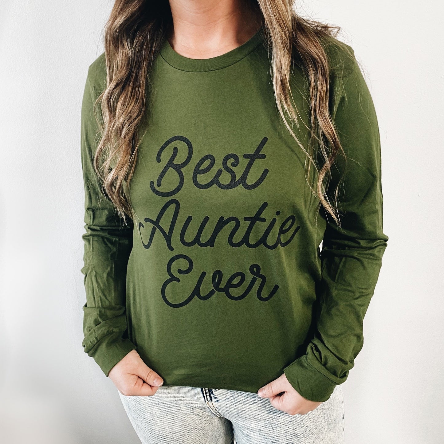 A person wears the Best Auntie Ever • Olive Long Sleeve, a unisex shirt with bold black script, standing against a plain white background.