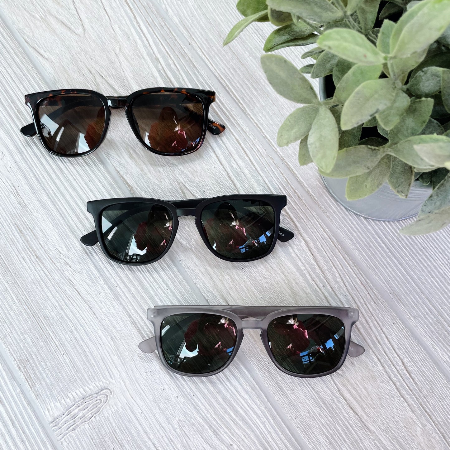 Three pairs of Simon Sunglasses (adult size, UV400, dark lenses) in tortoiseshell, black, and gray are displayed side by side on a light wooden surface next to a green potted plant. More colors available!.