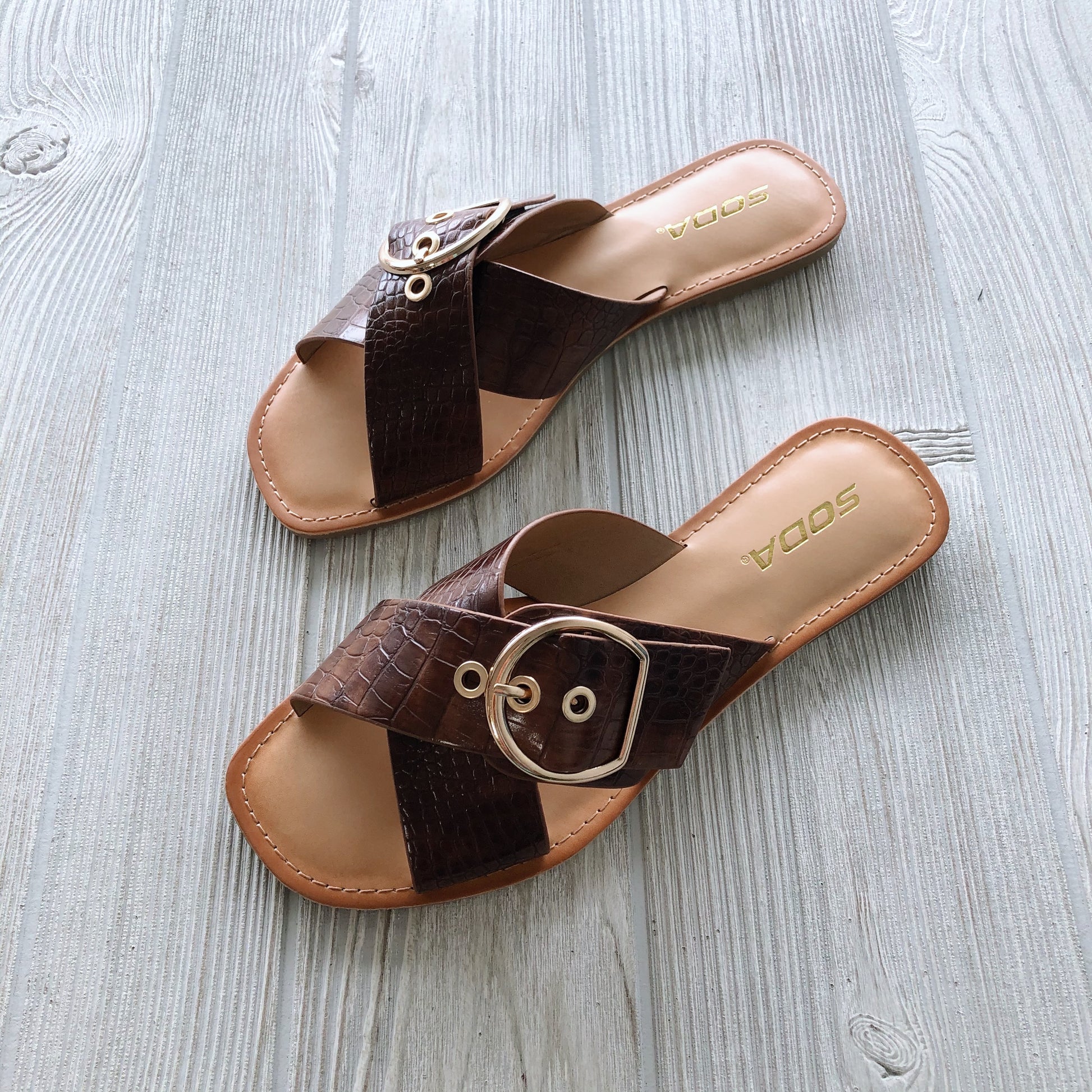 A pair of cognac Buckle Sandal with crisscross faux crocodile straps, large gold buckle, and gold SODA branding on tan insoles, shown on a light wooden floor. Available in Cognac or Black!.