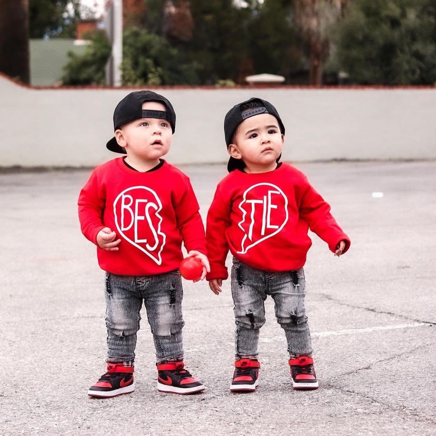 Two toddlers stand outdoors in matching red Bestie Pullover • Red sweatshirts, wearing backwards caps and ripped jeans. Holding hands, they look up—perfect best friends in stylish, unisex youth pullovers.
