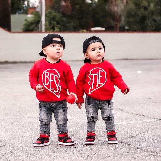 Two toddlers stand outdoors in matching red Bestie Pullover • Red sweatshirts, wearing backwards caps and ripped jeans. Holding hands, they look up—perfect best friends in stylish, unisex youth pullovers.