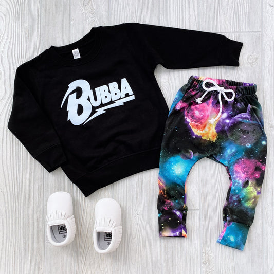 A stylish rocker outfit with the black Bowie Bubba • Kids Sweatshirt, galaxy-print pants with a white drawstring, and white slip-on shoes—arranged on a light wood floor. For longer wear, consider sizing up in sweatshirts!.