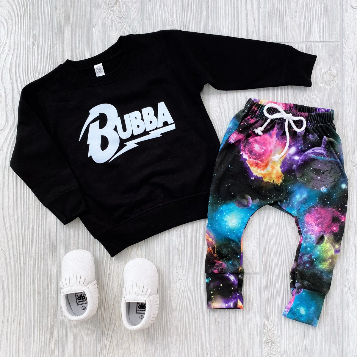 A stylish rocker outfit with the black Bowie Bubba • Kids Sweatshirt, galaxy-print pants with a white drawstring, and white slip-on shoes—arranged on a light wood floor. For longer wear, consider sizing up in sweatshirts!.