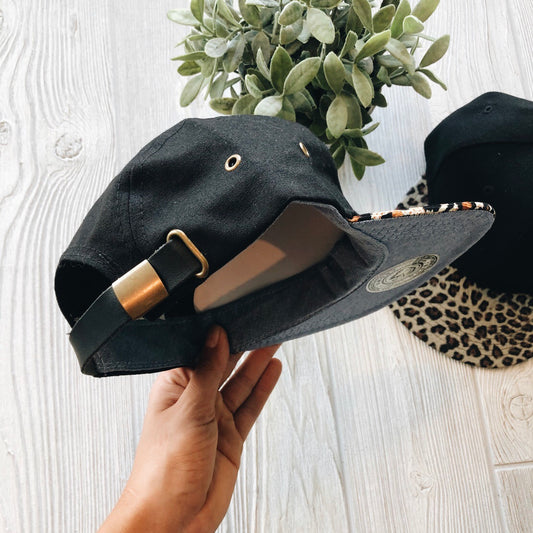 A hand holds the Mommy & Me • Cheetah Hat, a black cap with a brass buckle and leopard print under the brim and an adjustable strap. Nearby, a child-sized version with a full cheetah print brim sits on a light wooden surface by a green plant.