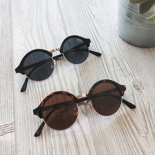Two pairs of Luna Sunglasses • Adult with UV400 dark lenses are on a light wooden surface—one with black frames, one tortoiseshell. A metal plant pot appears in the upper right corner.