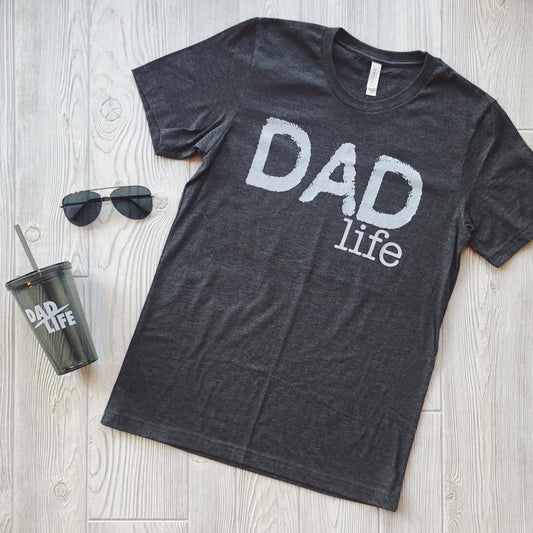 The black Dad Life Tee, made from soft combed and ring-spun cotton with DAD life in white, is shown on a light wood floor next to dark sunglasses and a tumbler cup with the matching print.