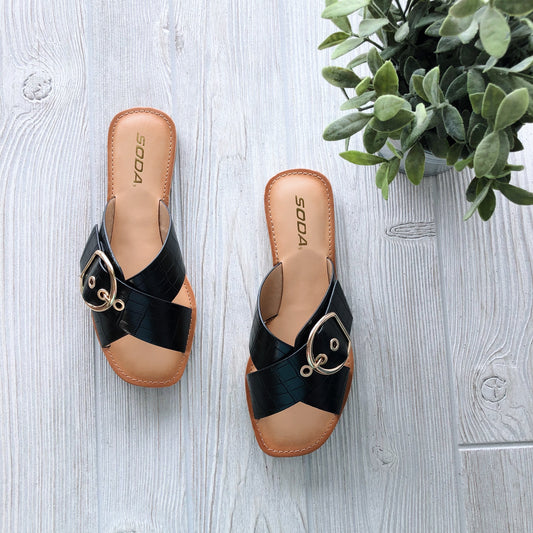 A pair of Buckle Sandal in black with gold buckles and faux crocodile print sits on a light wood floor beside a green leafy plant. The SODA label appears on the insole. Available in Cognac or Black!.