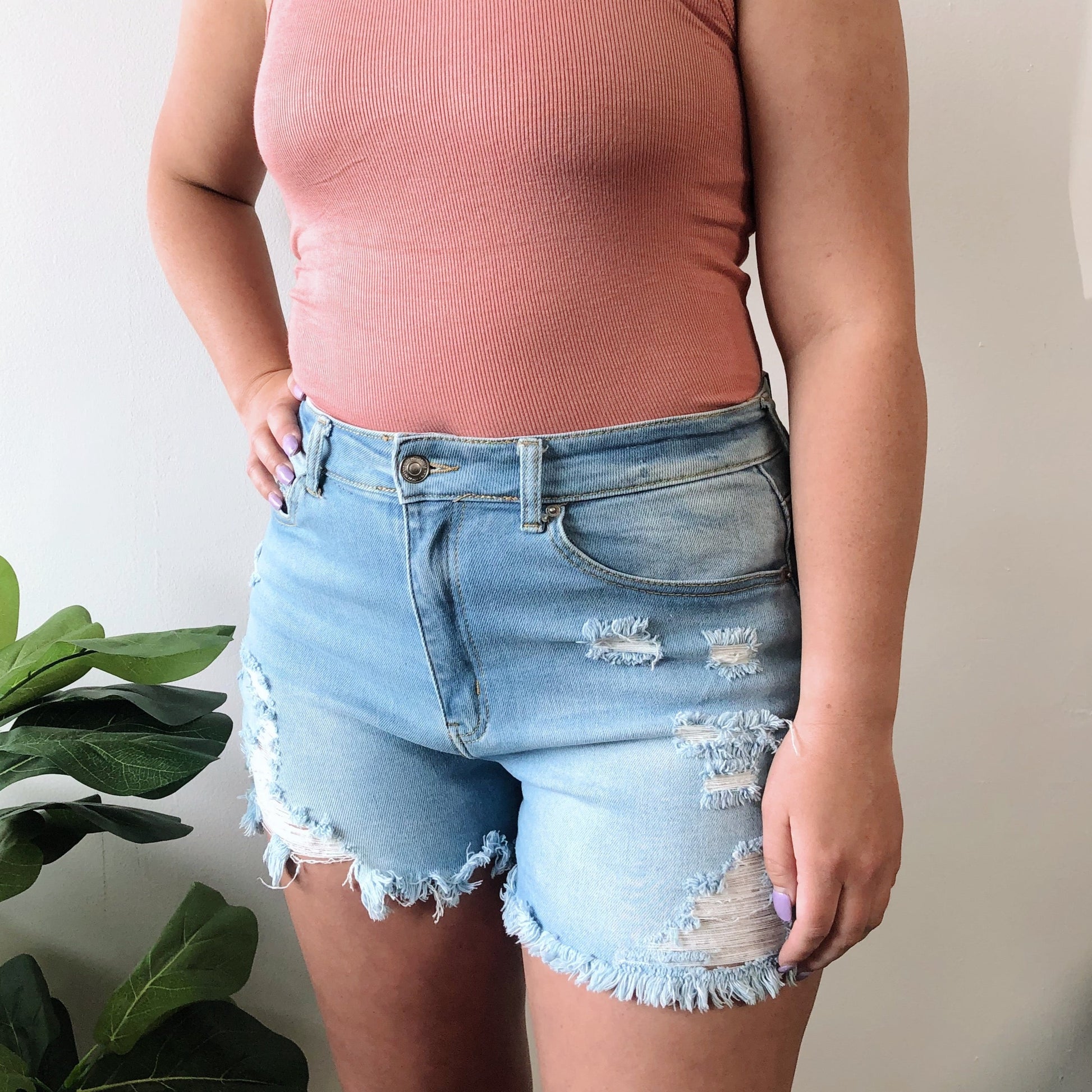 A person wearing a sleeveless, ribbed pink top and Beck Denim Shorts • Light Wash stands near a leafy green plant against a plain light-colored wall.