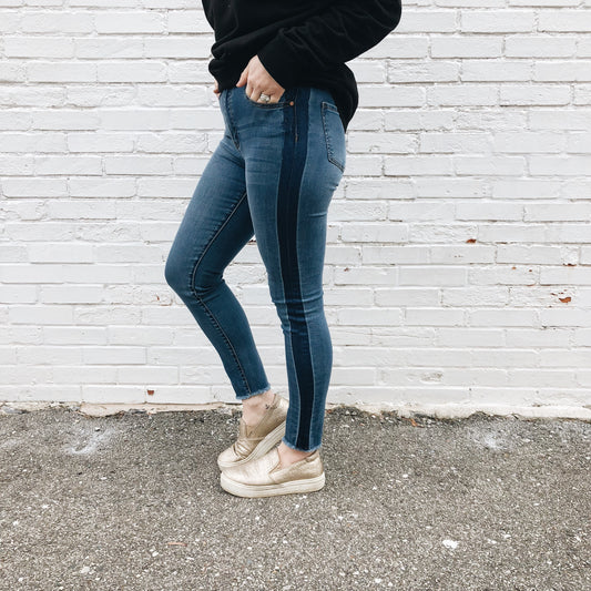 Only the lower half of a person is visible, wearing Alie Jeggings SIZE 0 & 3—blue super-stretch skinny jeans with a darker side stripe—paired with a black sweater and metallic gold slip-on shoes, standing against a white brick wall.