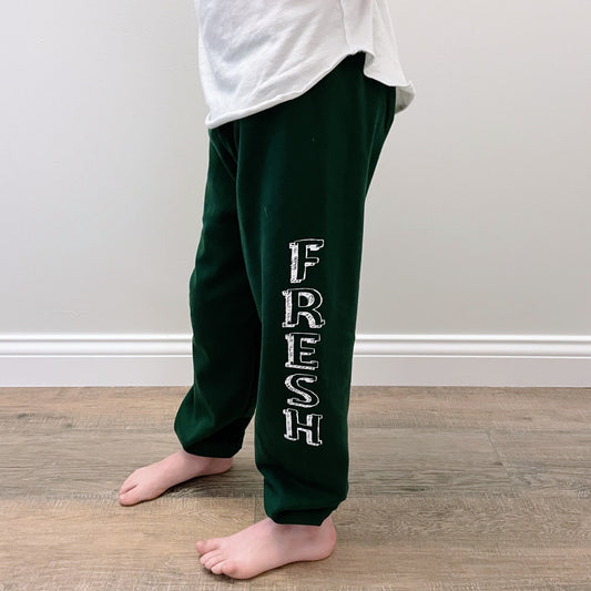 A child stands barefoot on a wooden floor, wearing Fresh Sweatpants • Kids in dark green with FRESH printed vertically in white on one leg, paired with a white t-shirt.