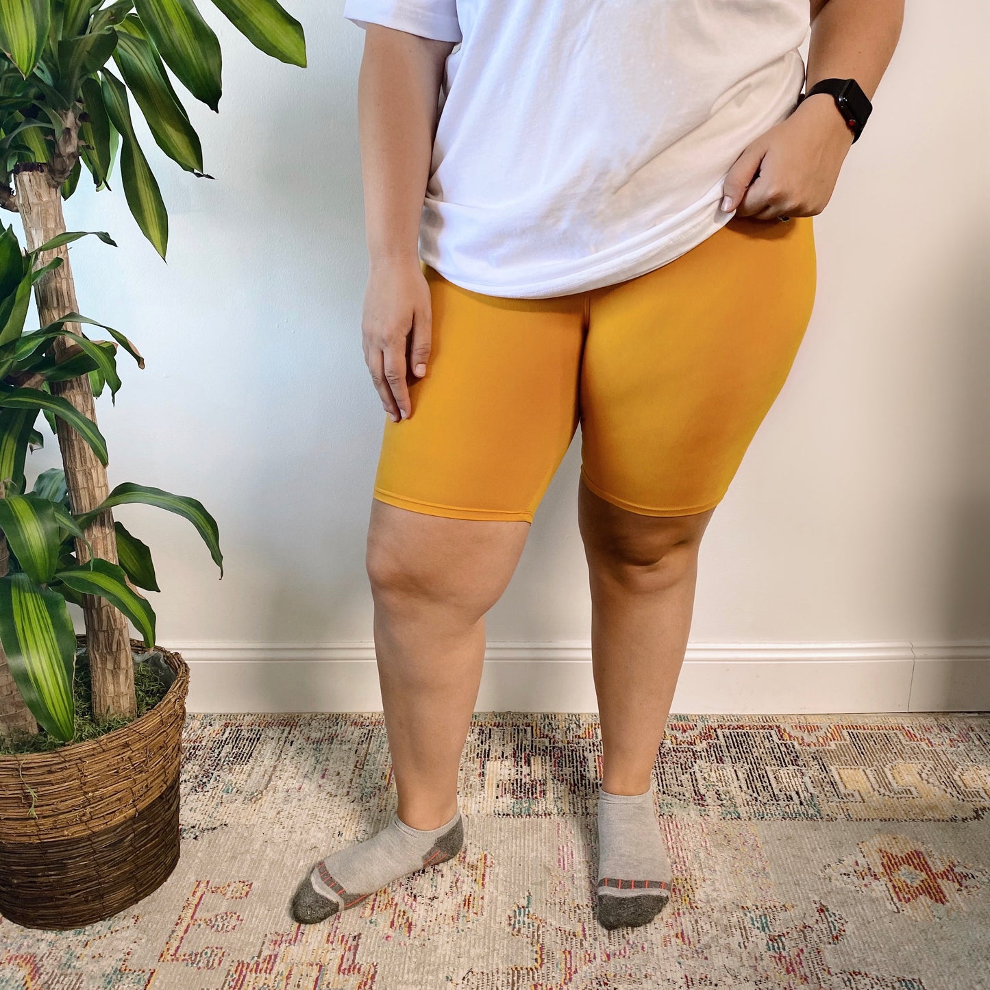 A person stands indoors next to a potted plant on a patterned rug, wearing Mustard • Plus Biker Shorts, a white t-shirt, and gray socks. Their face is not visible.