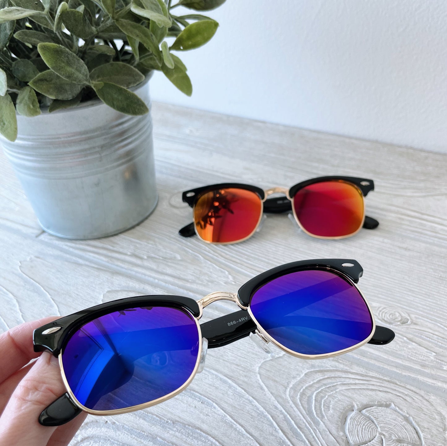 A hand holds Parker • Adult Sunglasses with blue mirrored lenses; another pair with red-orange mirrored lenses rests on a light wooden surface beside a potted plant in a metallic container. Final Sale.