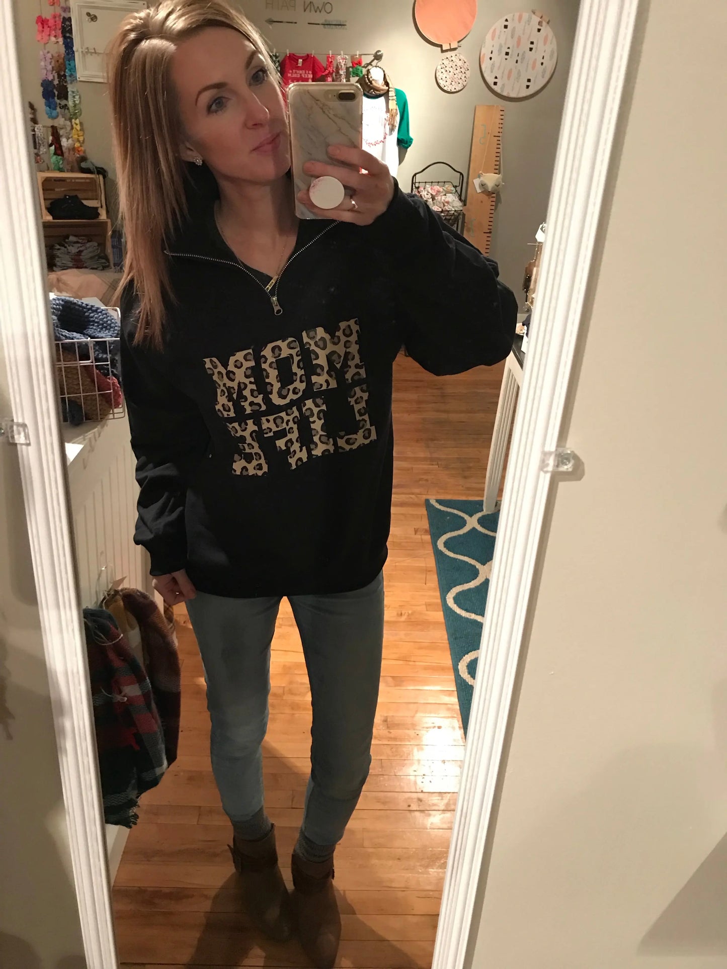 A woman takes a mirror selfie in a cozy room, wearing the Mom Life Cheetah • Quarter Zip with bold cheetah print, light blue jeans, and brown boots. The space has wooden floors, a blue rug, and craft supplies visible in the background.