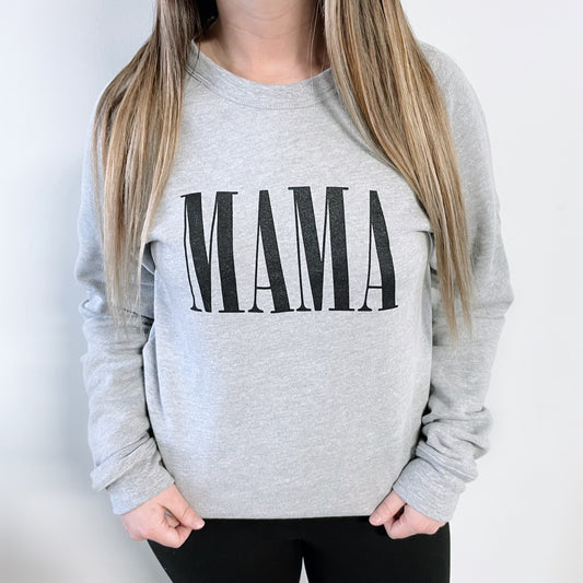 A person with long, straight blonde hair wears the gray MAMA Bella Pullover featuring bold black lettering, paired with black pants. They stand against a plain white background with hands in their pockets.
