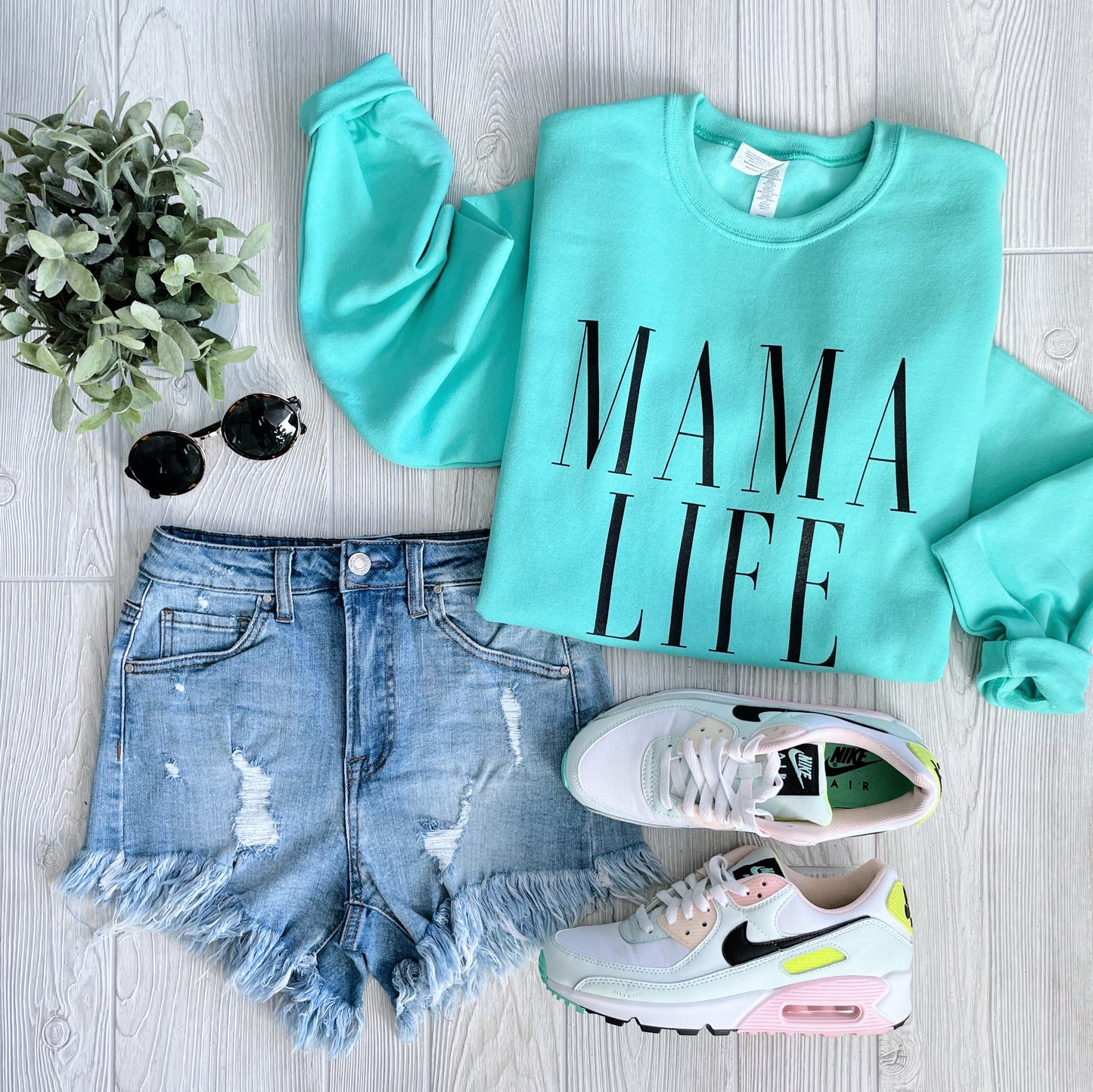 Flat lay of the MAMA LIFE • Mint Pullover with unisex fit, paired with distressed denim shorts, white sneakers with pink and yellow accents, black sunglasses, and a small green potted plant on a light wood floor—ideal for cooler days.
