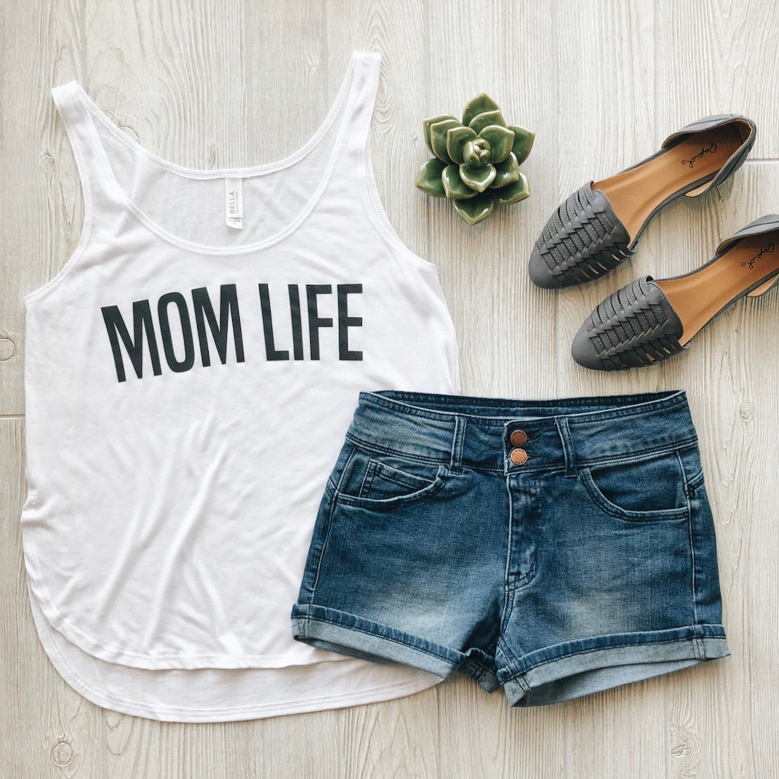 Flat lay of the MOM LIFE Block • Side Slit Tank in white, paired with blue denim shorts, gray woven slip-on shoes, and a small green succulent on a light wooden surface.