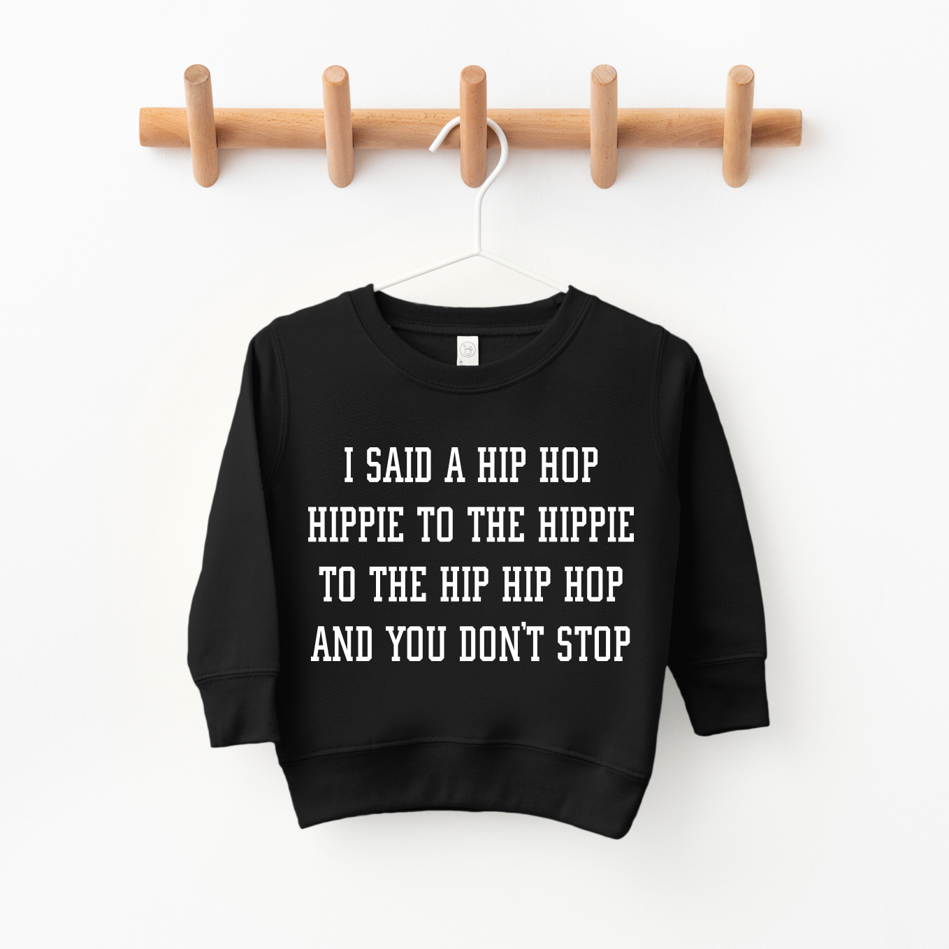 The Hip Hop Pullover, a black unisex sweatshirt with white text saying I SAID A HIP HOP HIPPIE TO THE HIPPIE TO THE HIP HIP HOP AND YOU DONT STOP, hangs on a wooden wall rack—ideal for kids or sized up for a comfy fit.