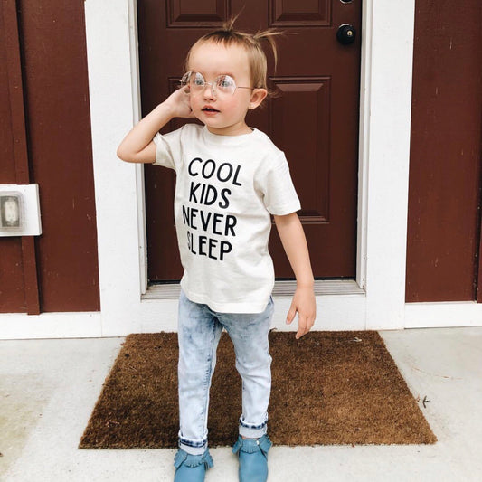 A young hipster babe with glasses stands on a doormat in front of a brown door, wearing a COOL KIDS NEVER SLEEP shirt and Lennon Sunnies • Kids—ready to swap style any day.