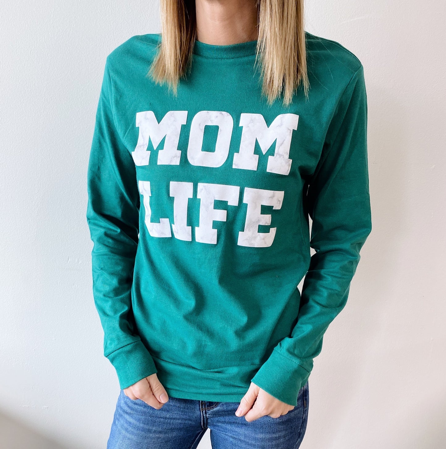 Someone stands against a plain white background wearing the Mom Marble • Emerald Long Sleeve shirt, which features large white MOM LIFE lettering, paired with blue jeans.