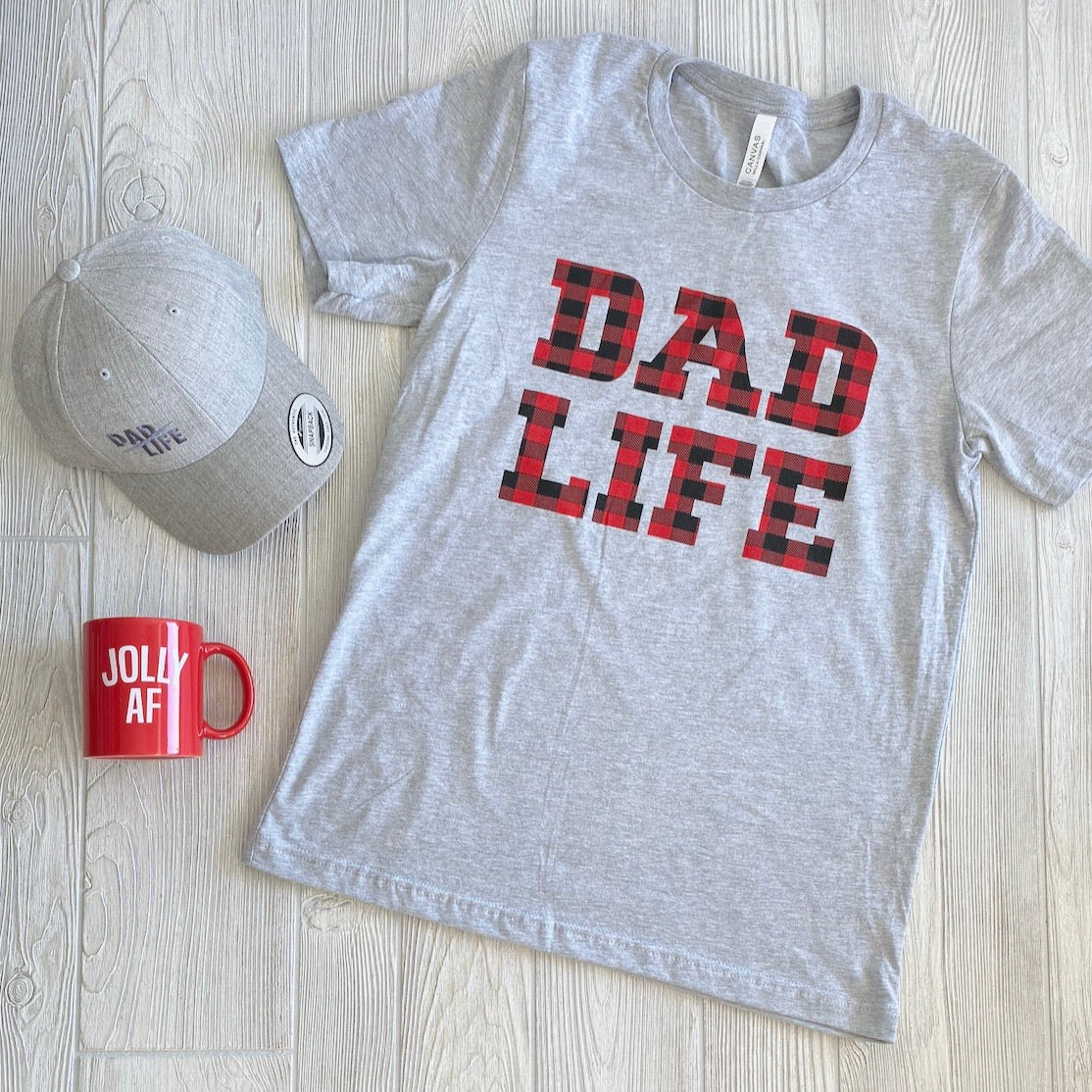 The Dad Life • Buffalo Plaid gray unisex t-shirt, made in Michigan with red and black plaid DAD LIFE letters, is shown on a light wood surface beside a matching gray cap and a red mug with white JOLLY AF text.