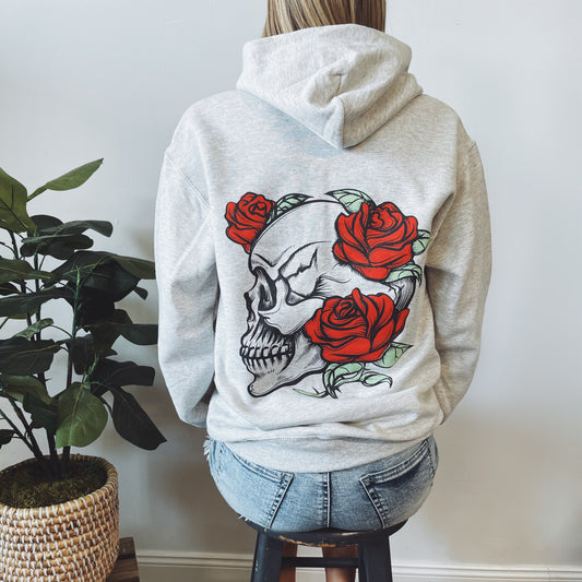 Someone sits on a stool next to a potted plant, facing away from the camera, wearing the Mother Skull • Oatmeal Hoodie—a gray unisex hoodie featuring a large skull and red roses graphic on the back, made from USA grown cotton.