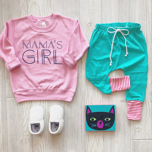 A flat lay displays a light pink Mamas Girl Pullover, teal pants with pink striped cuffs, white slip-on shoes, and a cat-illustrated book on a light wood floor.