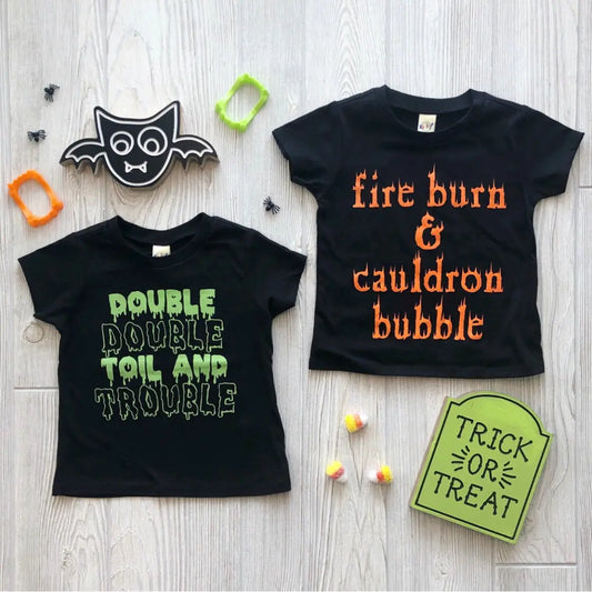 Two black unisex Cauldron Bubble kids tees for Halloween on a light wood surface. One says “DOUBLE DOUBLE TOIL AND TROUBLE” in green, the other “fire burn & cauldron bubble” in orange, surrounded by props and candy. Fun matching design.