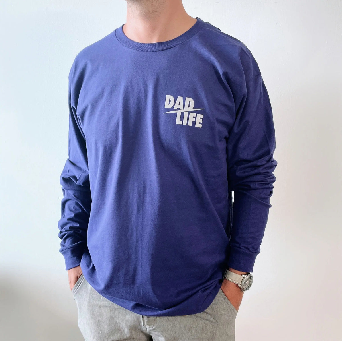 A man stands against a plain light background with his hands in his pockets, wearing the Dad Life Long Sleeve—a blue shirt featuring DAD LIFE in white on the chest and made from soft ring-spun cotton.