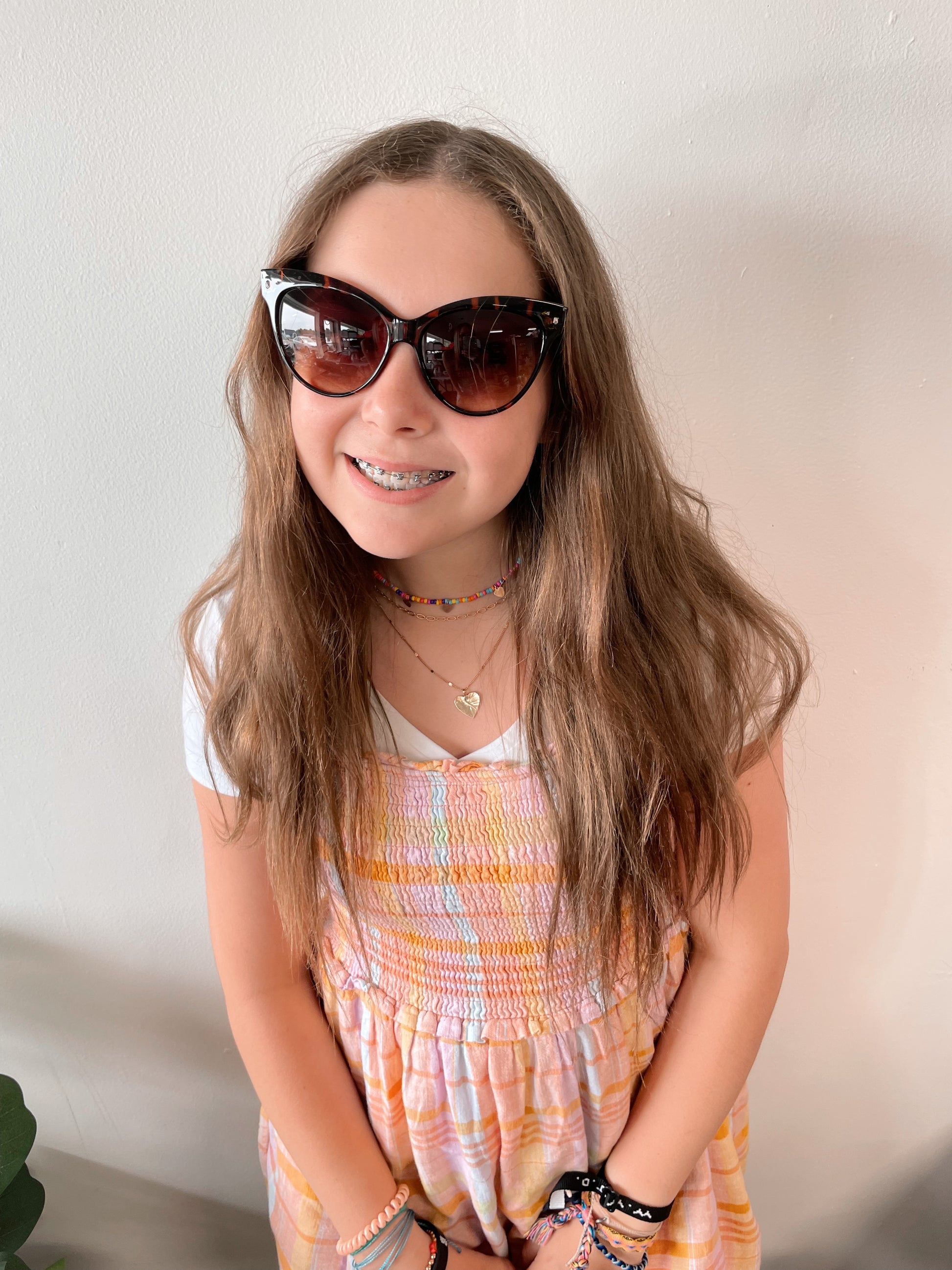 A smiling girl with long brown hair wears Sorya Sunglasses • Adult, featuring cat-eye frames and UV400 protection, along with a white shirt under a pastel pink and yellow dress, plus bracelets and necklaces, standing by a light-colored wall.