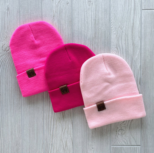 Three Valentines Collection Beanies in bright, hot, and light pink are displayed on a light wood surface. Each has a folded brim, front leather logo patch, and comes in one size for cozy style and comfort.