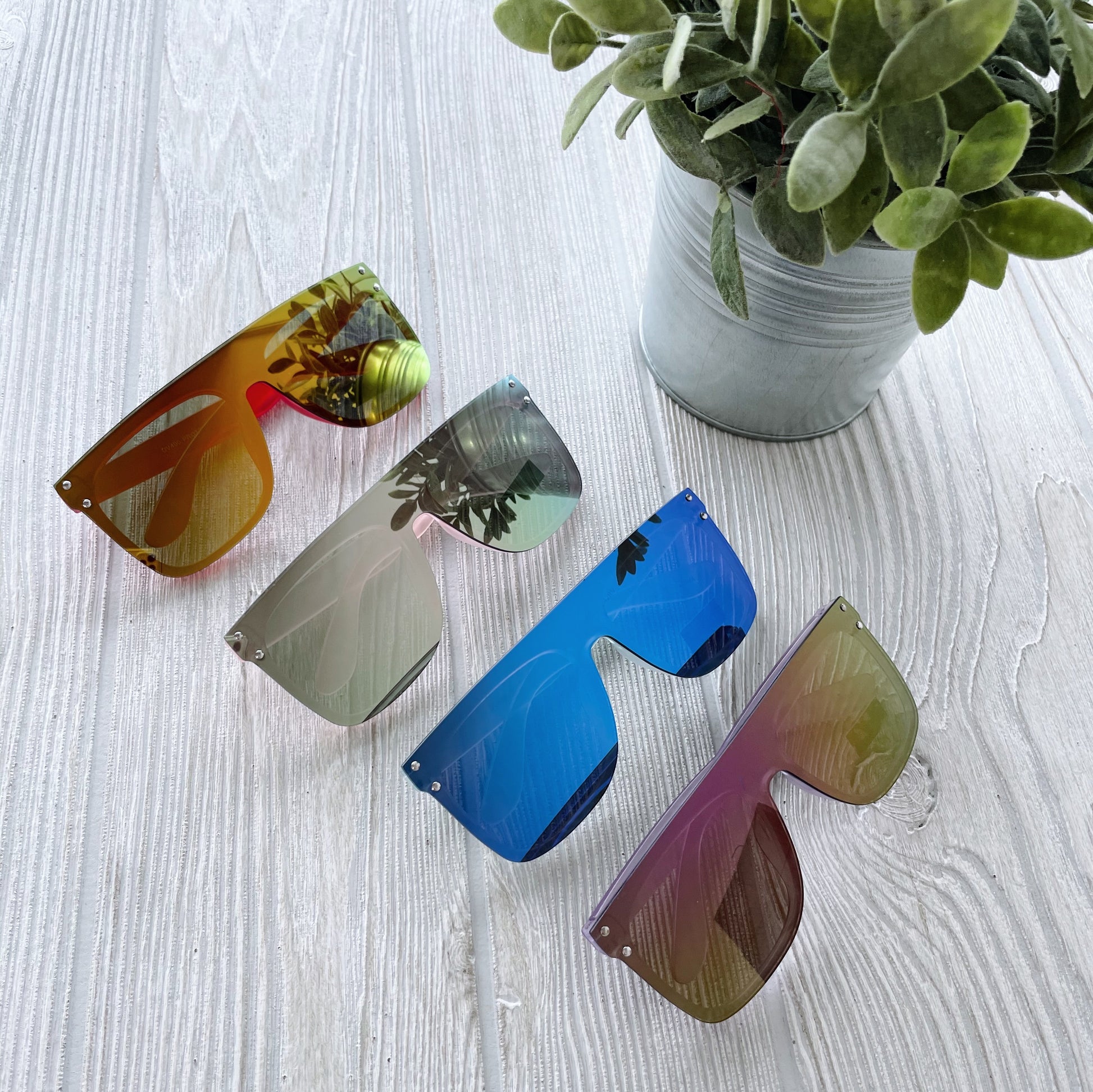 Four pairs of Arden Mirrored Sunnies • Kids, featuring vibrant colors and UV protection, are displayed on a light wooden surface beside a small green potted plant in a metal container.