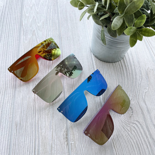 Four pairs of Arden Mirrored Sunnies • Kids, featuring vibrant colors and UV protection, are displayed on a light wooden surface beside a small green potted plant in a metal container.