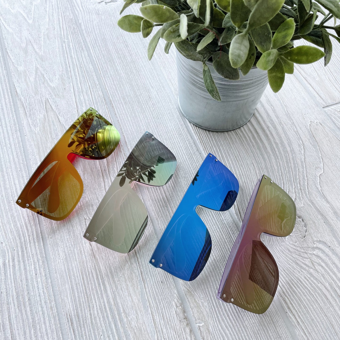 Four pairs of Arden Mirrored Sunnies • Kids, featuring UV-protected reflective lenses, are arranged on a light wooden surface beside a small potted plant, reflecting the nearby greenery.