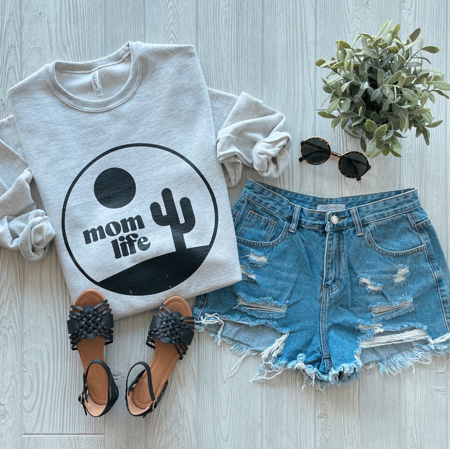 A flat lay of a Cactus Mom Life • Oatmeal Pullover featuring a cactus graphic, styled with ripped denim shorts, black braided sandals, round sunglasses, and a small potted plant on a light wood floor.