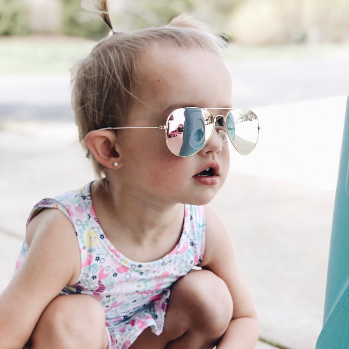A young child with light hair in a ponytail wears Aviator Sunnies • Kids and a colorful sleeveless dress while crouching outdoors on a sunny day.