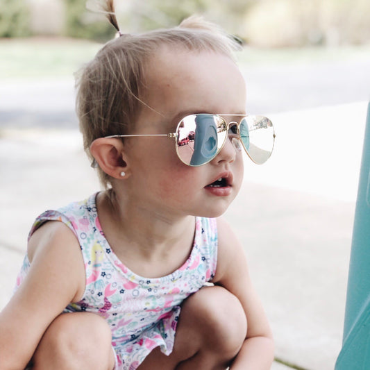 A young child with light hair in a ponytail wears Aviator Sunnies • Kids and a colorful sleeveless dress while crouching outdoors on a sunny day.