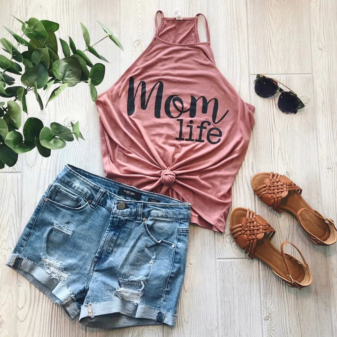 A flat lay of summer essentials: the Mom Life OG Mauve High Neck Tank, distressed denim shorts, tan sandals, dark sunglasses, and a green plant displayed on a light wooden floor.